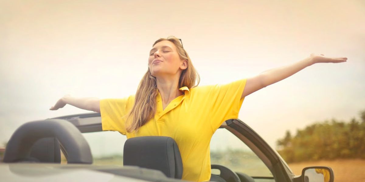 A cheerful woman in a yellow shirt enjoys freedom on a sunny drive in a convertible.
